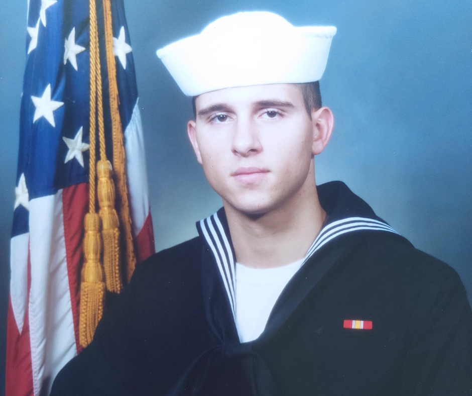 A young U.S. Navy service member in dress uniform stands in front of an American flag during a formal portrait.