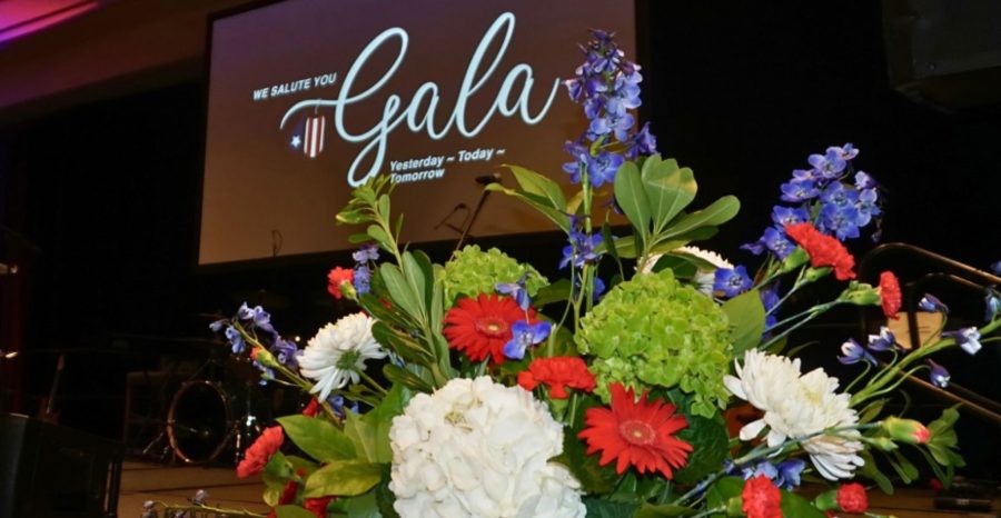 A large red, white, and blue floral arrangement sits in front of the stage at the USA Cares Gala, with the event screen and lighting in the background.