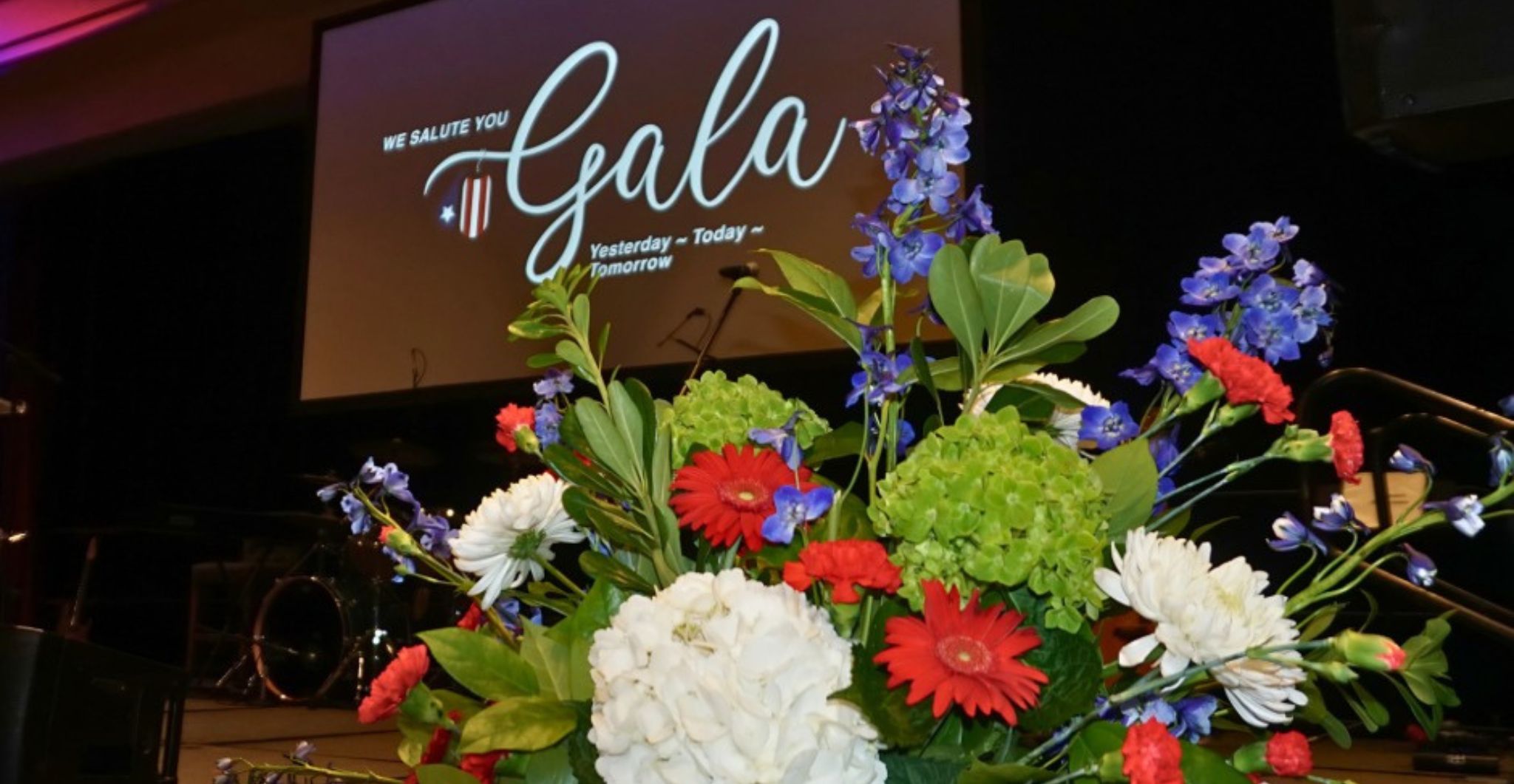 A large red, white, and blue floral arrangement sits in front of the stage at the USA Cares Gala, with the event screen and lighting in the background.